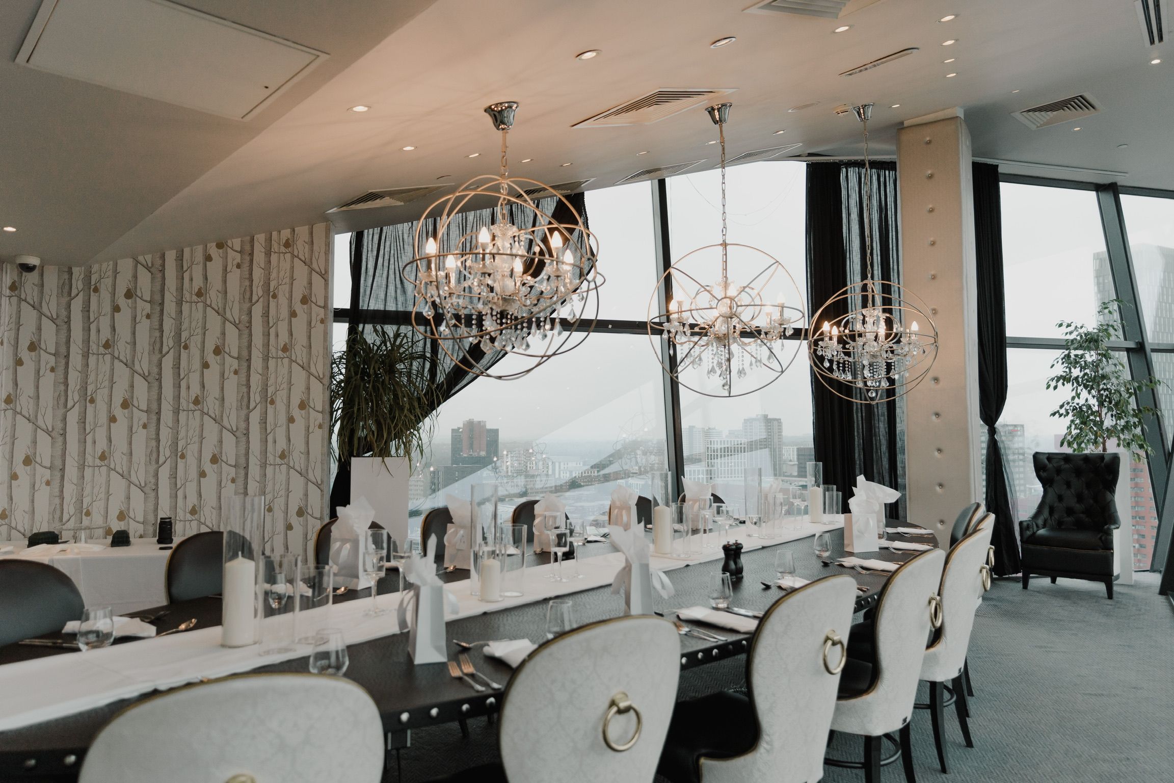 Wedding reception styled by LK's: a long table with a white runner, pillar candles in glass and favour bags beneath three crystal chandeliers, with city views through the windows